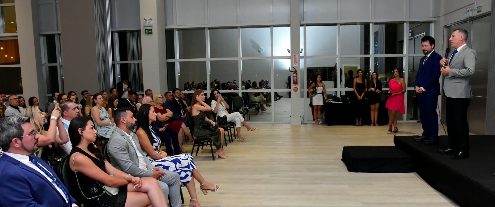 Convidados acompanham a fala do Dr. Anderson Macohin, ao microfone, ao lado de André Macohin, no palco, em 10 de janeiro de 2024.